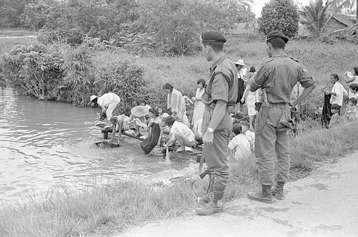 Armed soldiers stand guard in Sarawak in 1965 as a group of Chinese villagers take a communal bath. The authorities aim to protect the area from Indonesian raiders and to eliminate Communist influence.